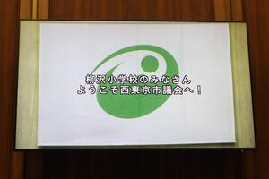 モニターに「柳沢小学校のみなさん　ようこそ西東京市議会へ！」が表示されている画像